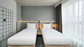 A modern hotel room with two single beds, a wooden headboard, a bedside table with a lamp, and a view of the city through sheer curtains. - Moxy Warsaw City (Warsaw)