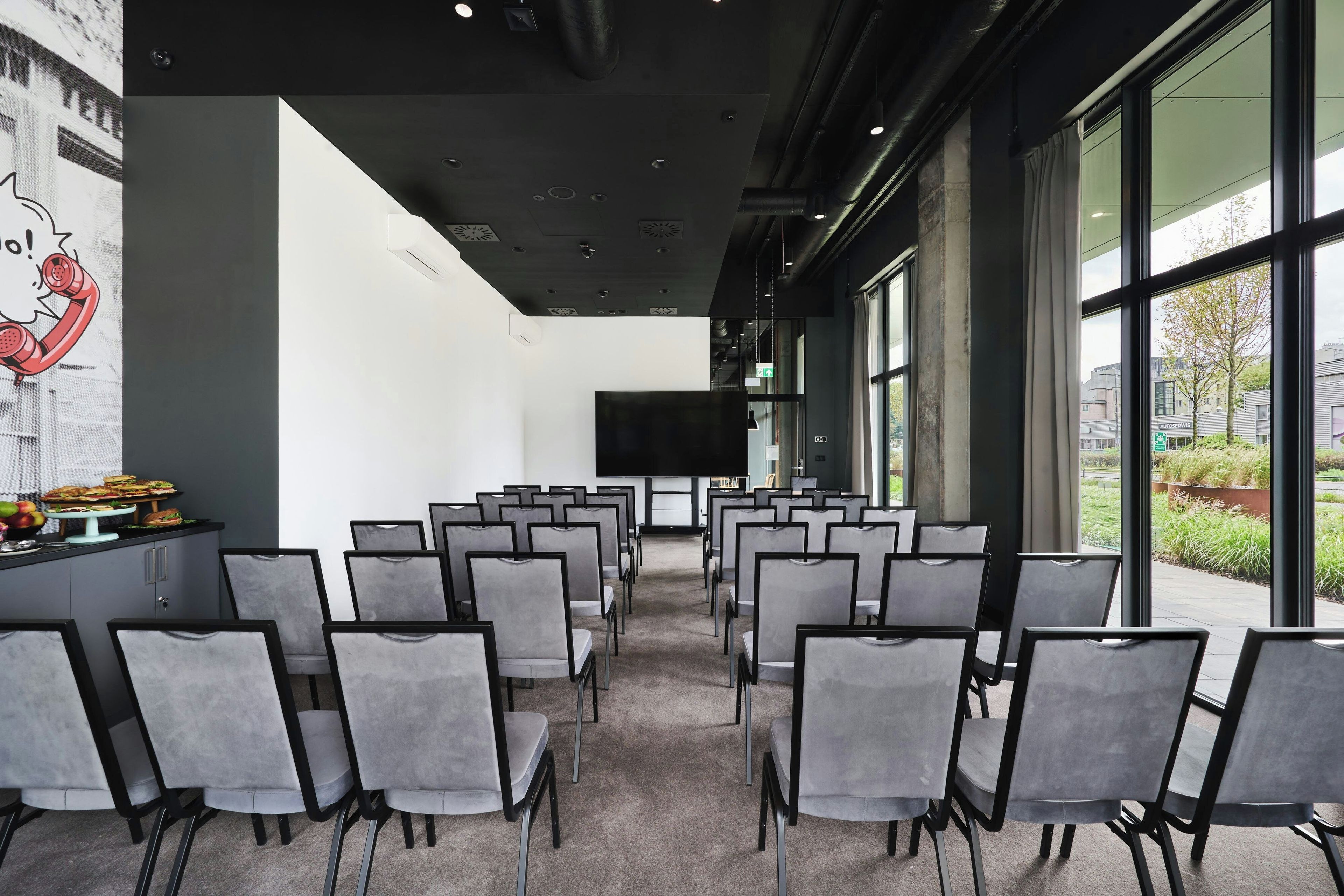 Un salón de conferencias con filas de sillas grises, un ventanal grande y un televisor de pantalla plana en la pared.