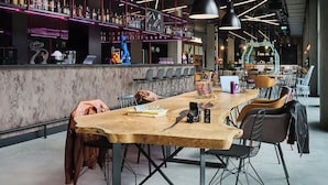 A modern bar area with a long wooden table, multiple chairs, and a bar counter with shelves of bottles. - Moxy Warsaw City (Warsaw)
