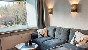 A modern living room with a grey sofa, two decorative pillows with deer motifs, a small round table with two mugs, and a window with a view of trees. - Apartment Elwi by Interhome (Hahnenklee)
