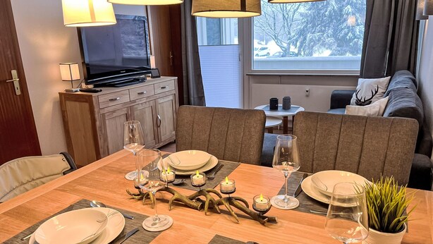 A dining area with a wooden table set for four, surrounded by cushioned chairs. A television is mounted on the wall, and a window offers a view of snow-covered trees. - Apartment Elwi by Interhome (Hahnenklee)