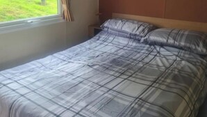 A bed with a plaid bedspread, a nightstand, a window with curtains, and a view of a green outdoor area. - 3 Bed Caravan - Sleeps 6 - Pets - Parking - Wifi (Borth)