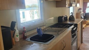 A compact kitchen with a gas stove, a sink, and a window with blinds. - 3 Bed Caravan - Sleeps 6 - Pets - Parking - Wifi (Borth)