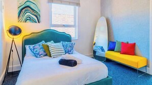 A bedroom with a bed, a bedside table, a lamp, a window, a sofa, and a wall art piece.
