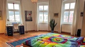 Une chambre à coucher avec une couvre-lit aux couleurs vives, un plancher en bois, un bureau avec un ordinateur et de grandes fenêtres avec des rideaux.