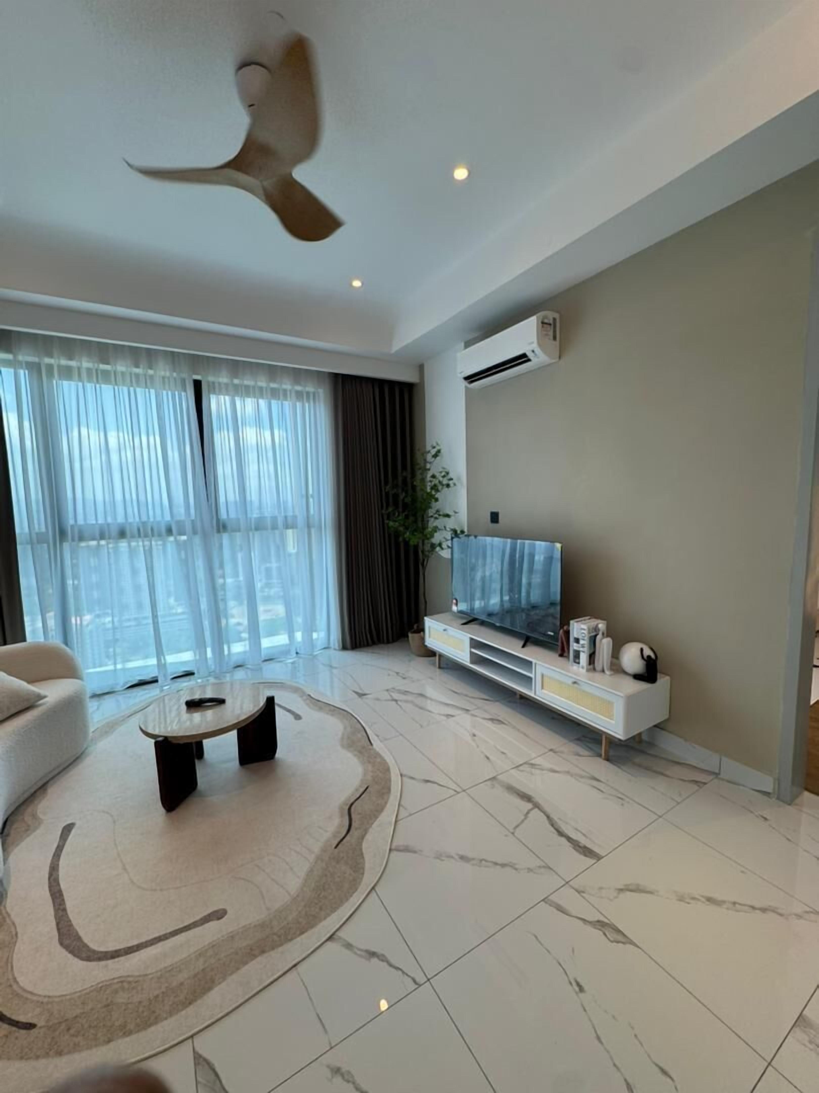 A modern living room with a large flat-screen TV, a ceiling fan, and a large window with sheer curtains.
