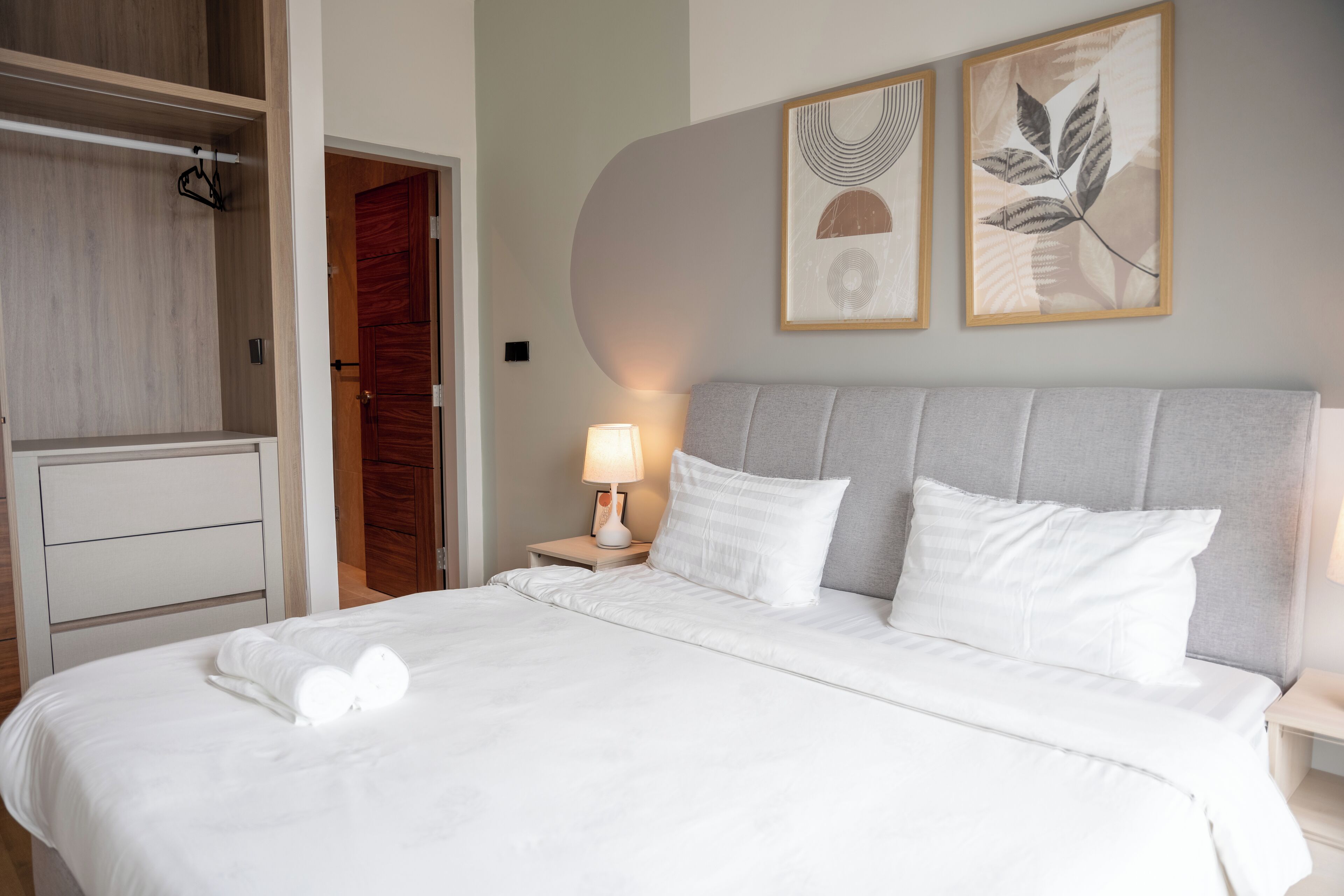Un letto rifatto con lenzuola e cuscini bianchi, una lampada da comodino e due quadri con foglie appese al muro.