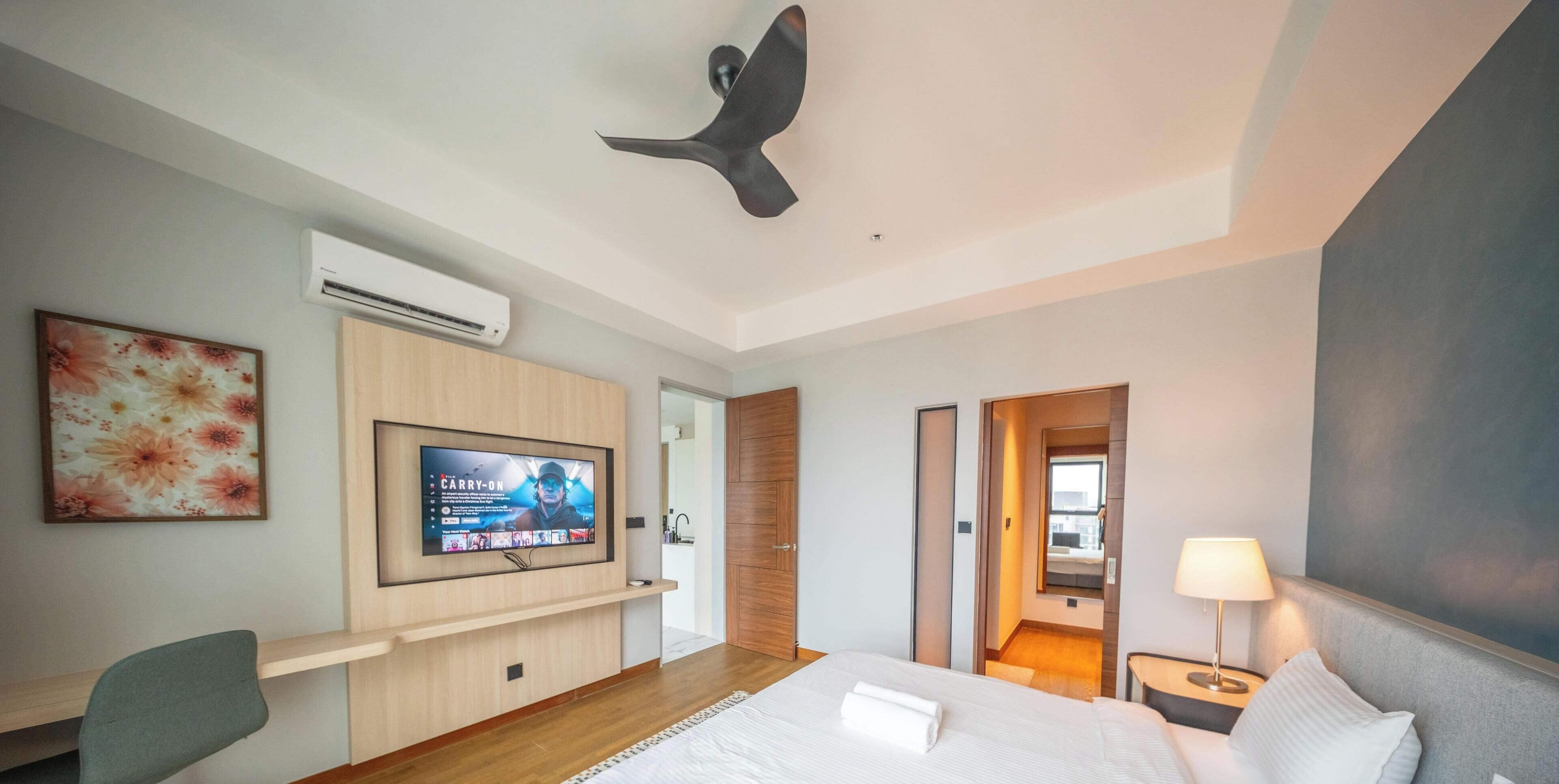 A modern hotel room with a large bed, a desk with a chair, a flat-screen TV mounted on the wall, and a ceiling fan.