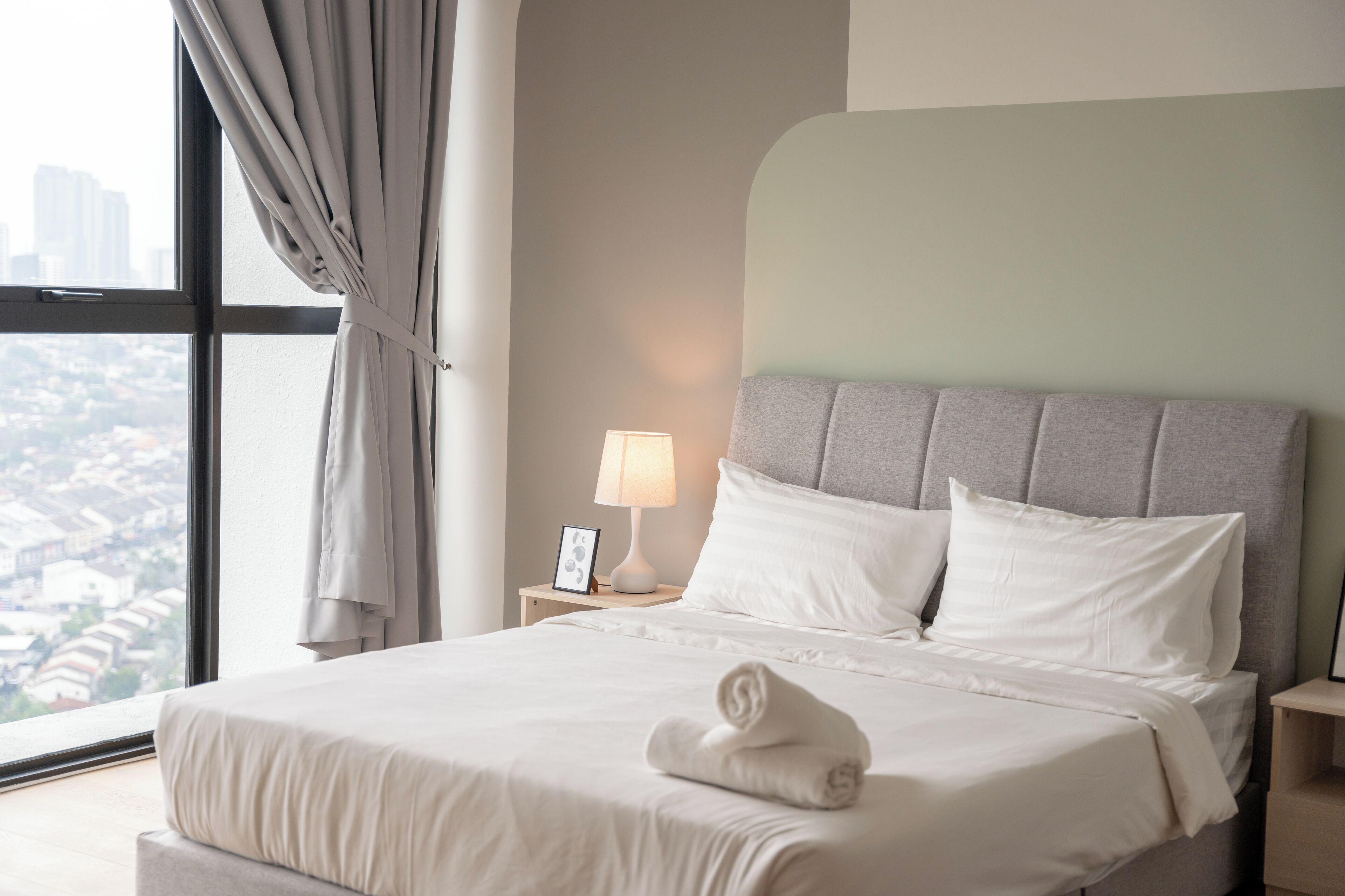 Una habitación de hotel con un ventanal grande, una cama con ropa blanca, un cabecero gris y una lámpara de noche.