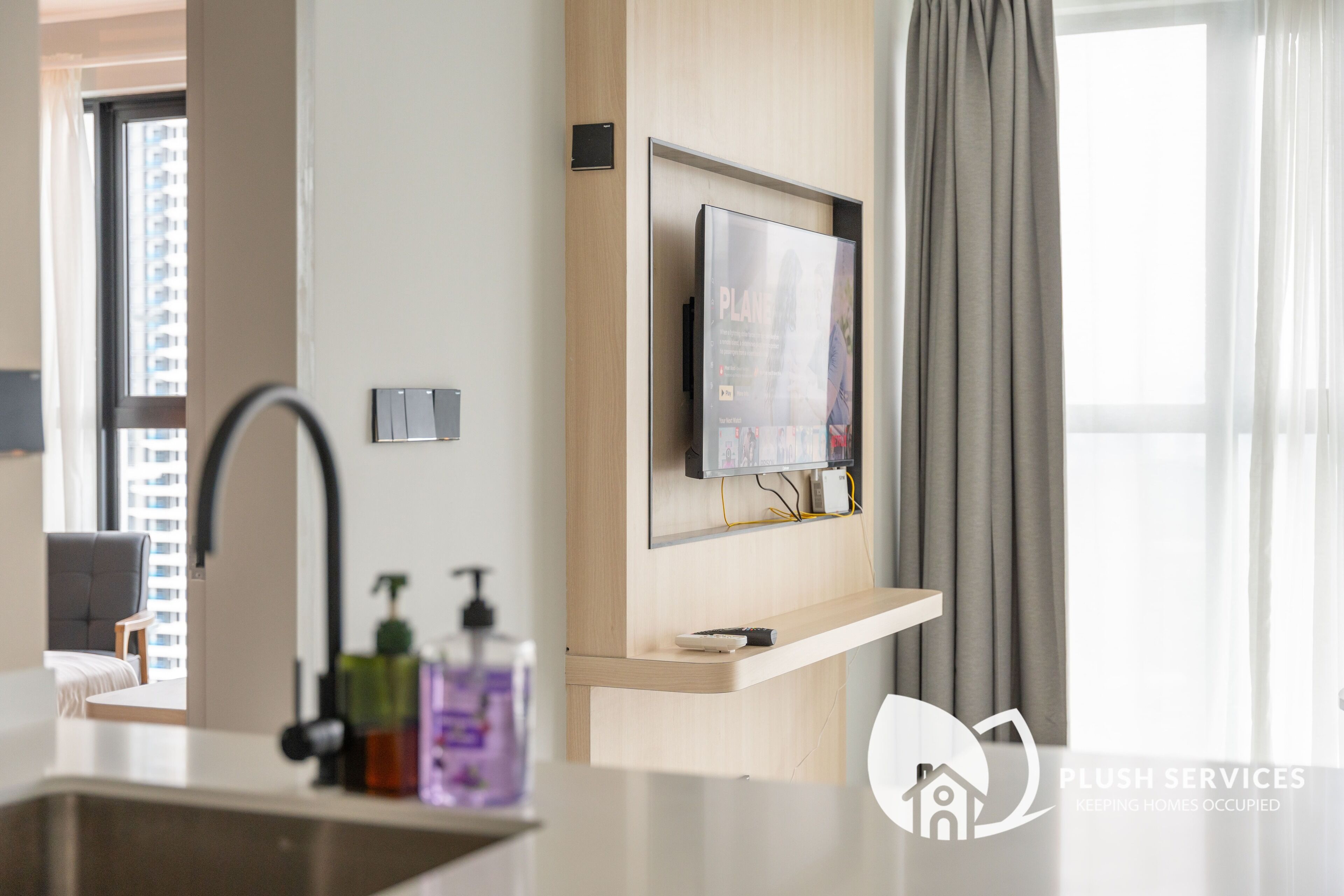 A modern hotel room with a mounted TV, a built-in shelf, a sink, and a window with curtains.
