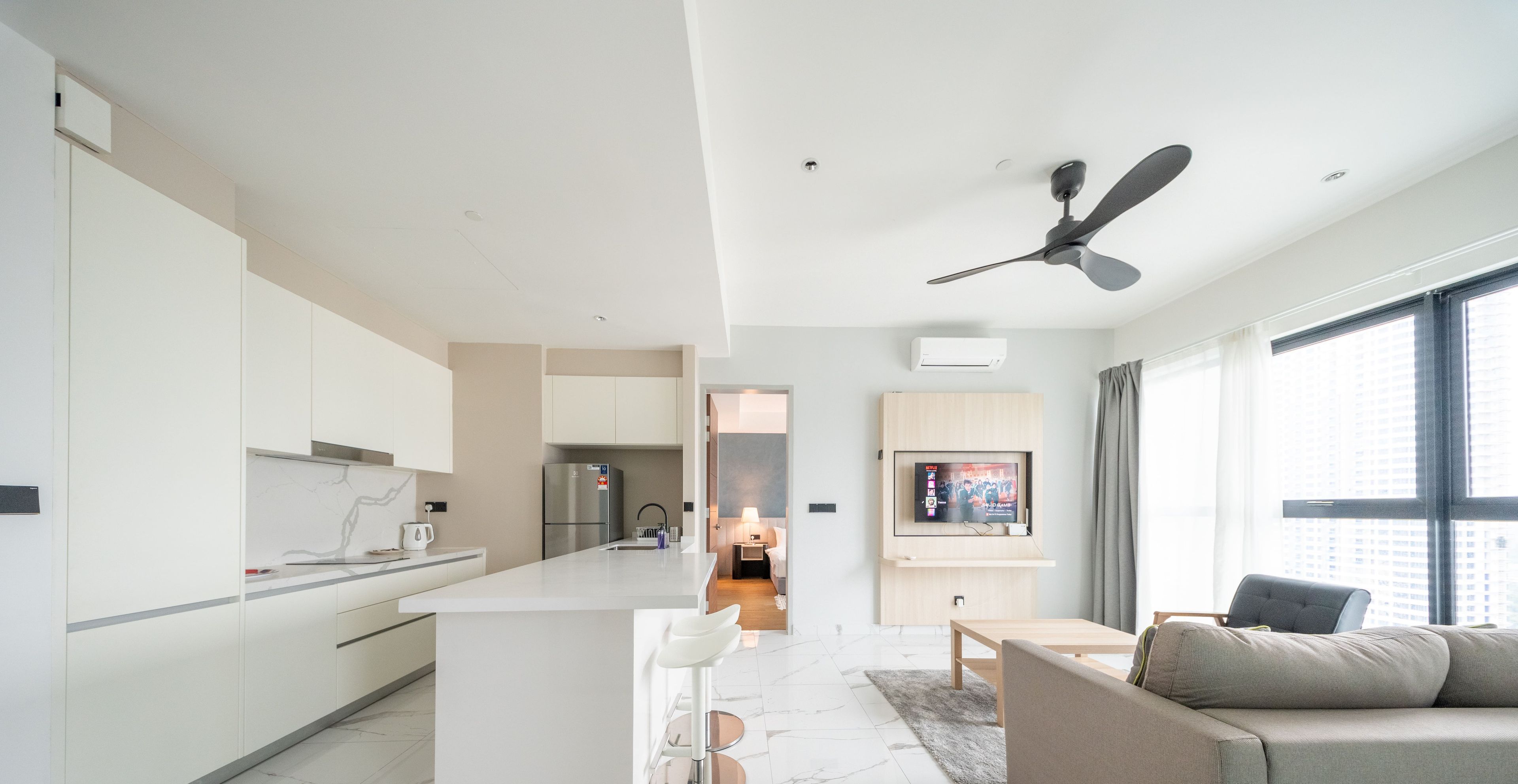 Una cocina moderna con armarios blancos, una isla central con taburetes de bar, un ventilador de techo y un amplio ventanal que proporciona luz natural.