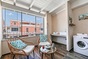 A balcony with wicker chairs, a small table with a book, a washing machine, and a view of buildings.