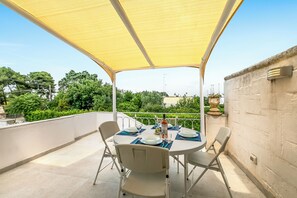 Une terrasse extérieure couverte, avec une table dressée pour deux personnes, entourée de chaises, et offrant une vue sur la verdure et les bâtiments en arrière-plan.