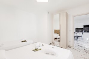 A modern hotel room with a large mirror, a bed, a small seating area, and a view into the kitchen area.