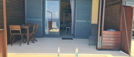 A room with a sliding glass door leading to a balcony with a view of the ocean. There are wooden chairs and a table on the balcony.
