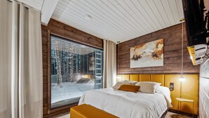 A modern bedroom with a large window, a bed with white bedding, a mustard-colored bench, and a wooden headboard.