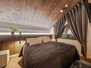 A bedroom with a wooden ceiling, a bed with a dark bedspread, a nightstand, a potted plant, and a window with curtains. - Levi star west a by Interhome (Kittilä)