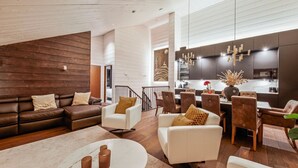 A modern living room with a brown leather sofa, white armchairs, and a round coffee table. A kitchen area with dark cabinets and a chandelier is visible in the background.
