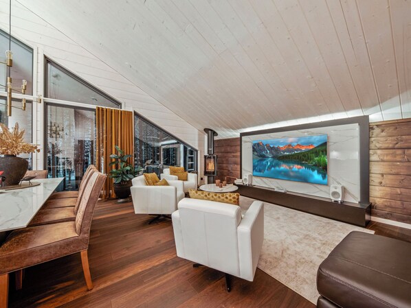 A modern living room with a fireplace, white sofas, a dining area with a glass table, and a large TV on the wall.