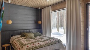 Un dormitorio con una cama grande, un banco, un taburete y vistas a un bosque nevado a través de una ventana.