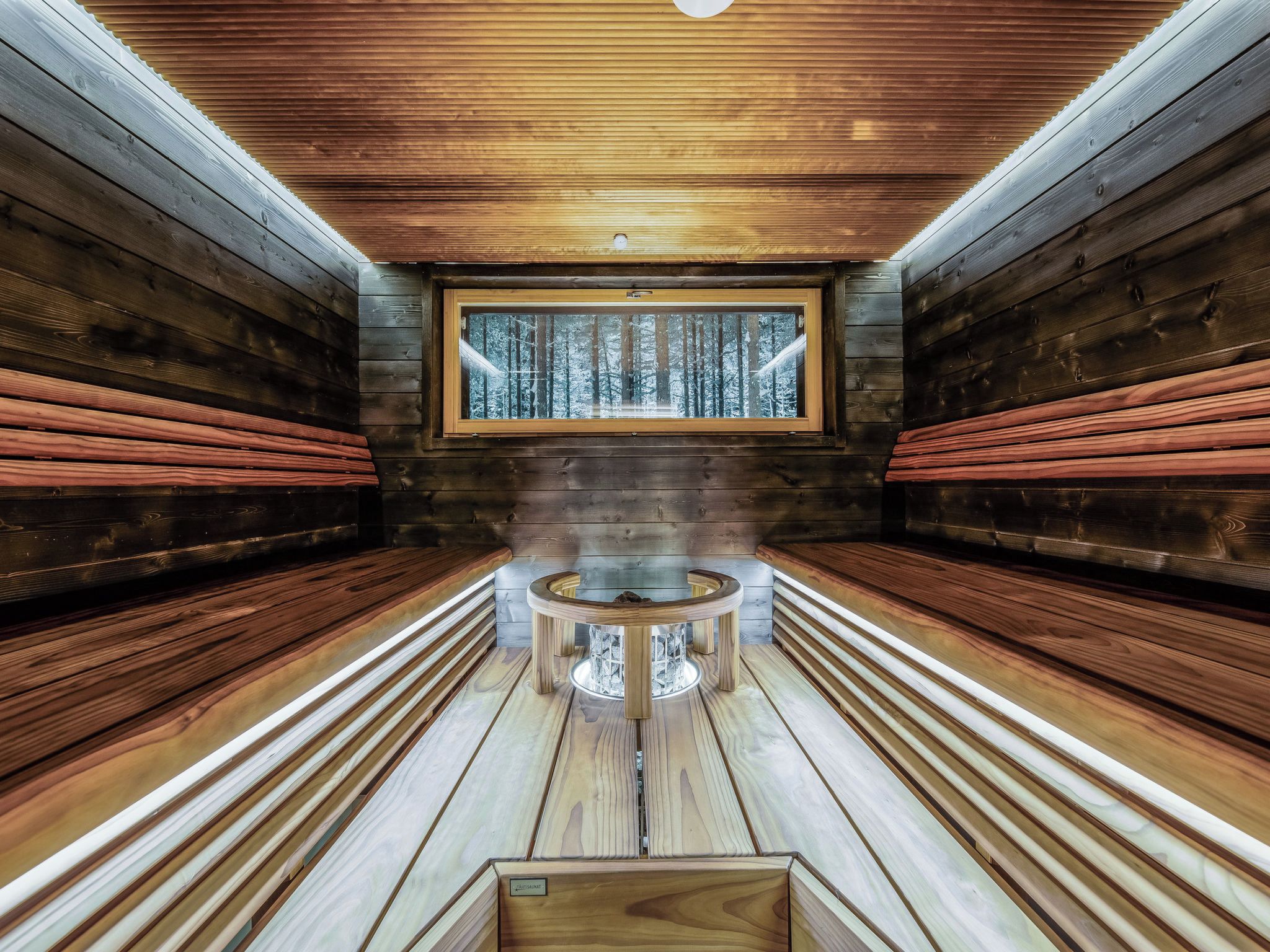 A sauna room with wooden benches, a central table, and a window with a snowy forest view.