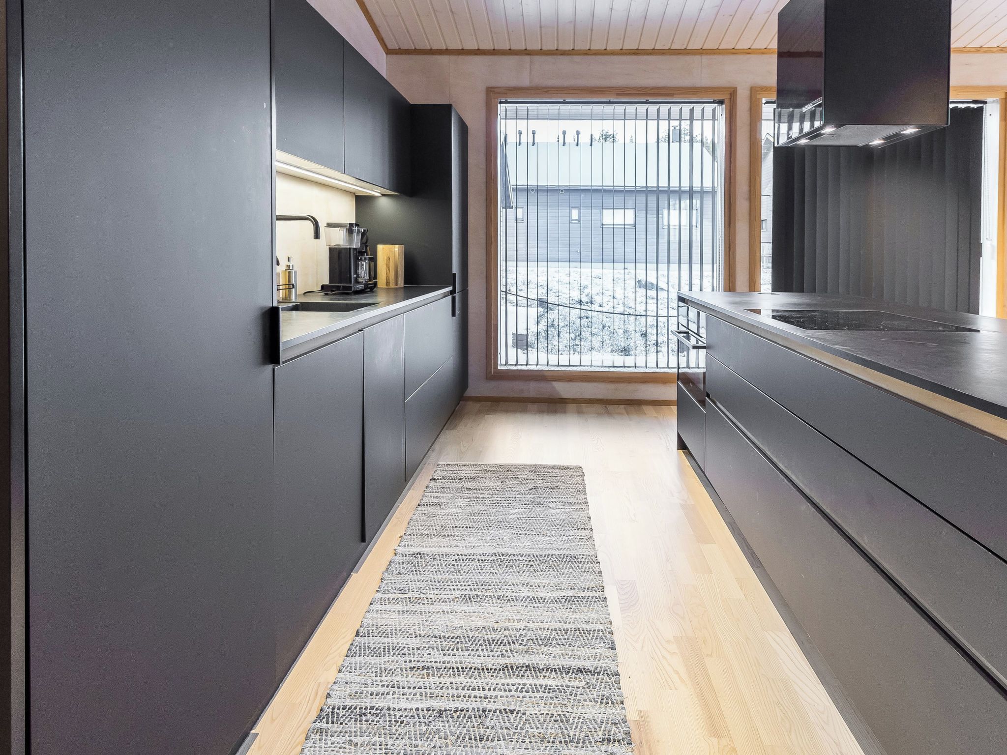A modern kitchen with dark cabinetry, a coffee machine, and a view through a window to a snowy outdoor area.
