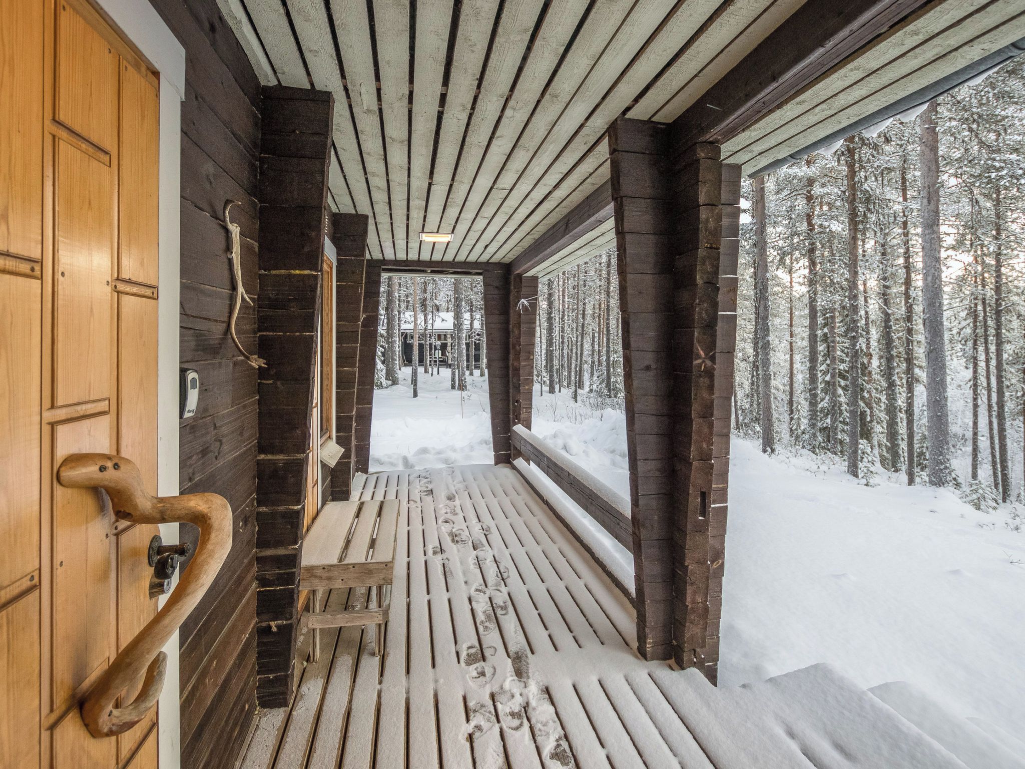 Ein Holzkorridor führt zu einer verschneiten Außenanlage, mit einer Bank und einer Tür mit einem markanten Griff.