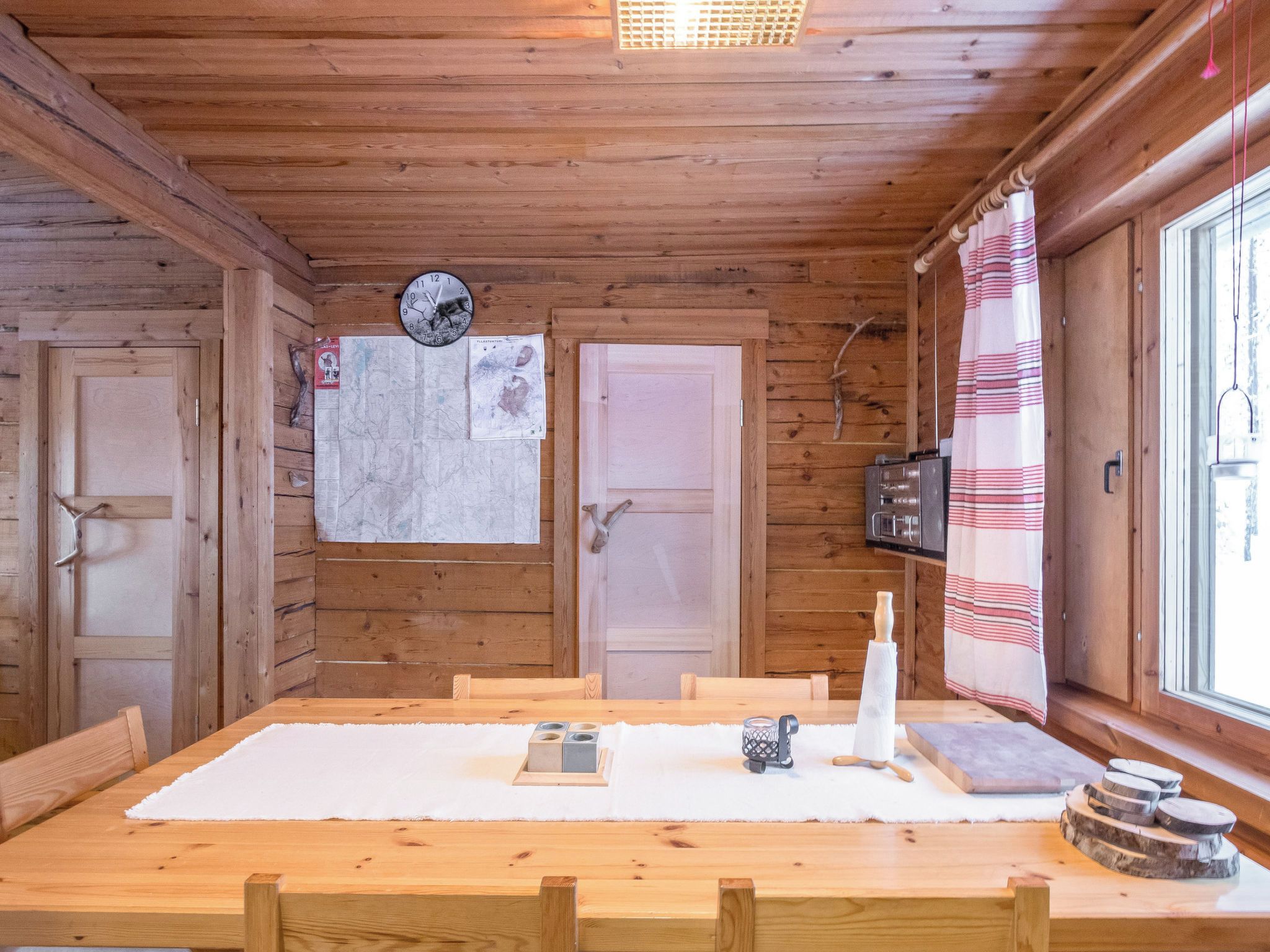 A wooden cabin interior with a large dining table, two doors, a wall clock, and a window with curtains.