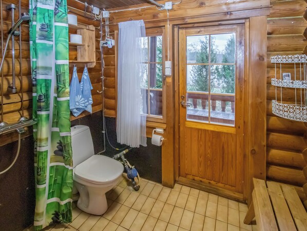 Baño de cabaña de madera con inodoro, ducha y ventana con vista a árboles.