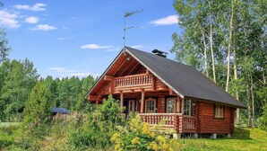 Hirsimökki, jossa on parveke, ja sen ympärillä on vehreää kasvillisuutta ja puita.