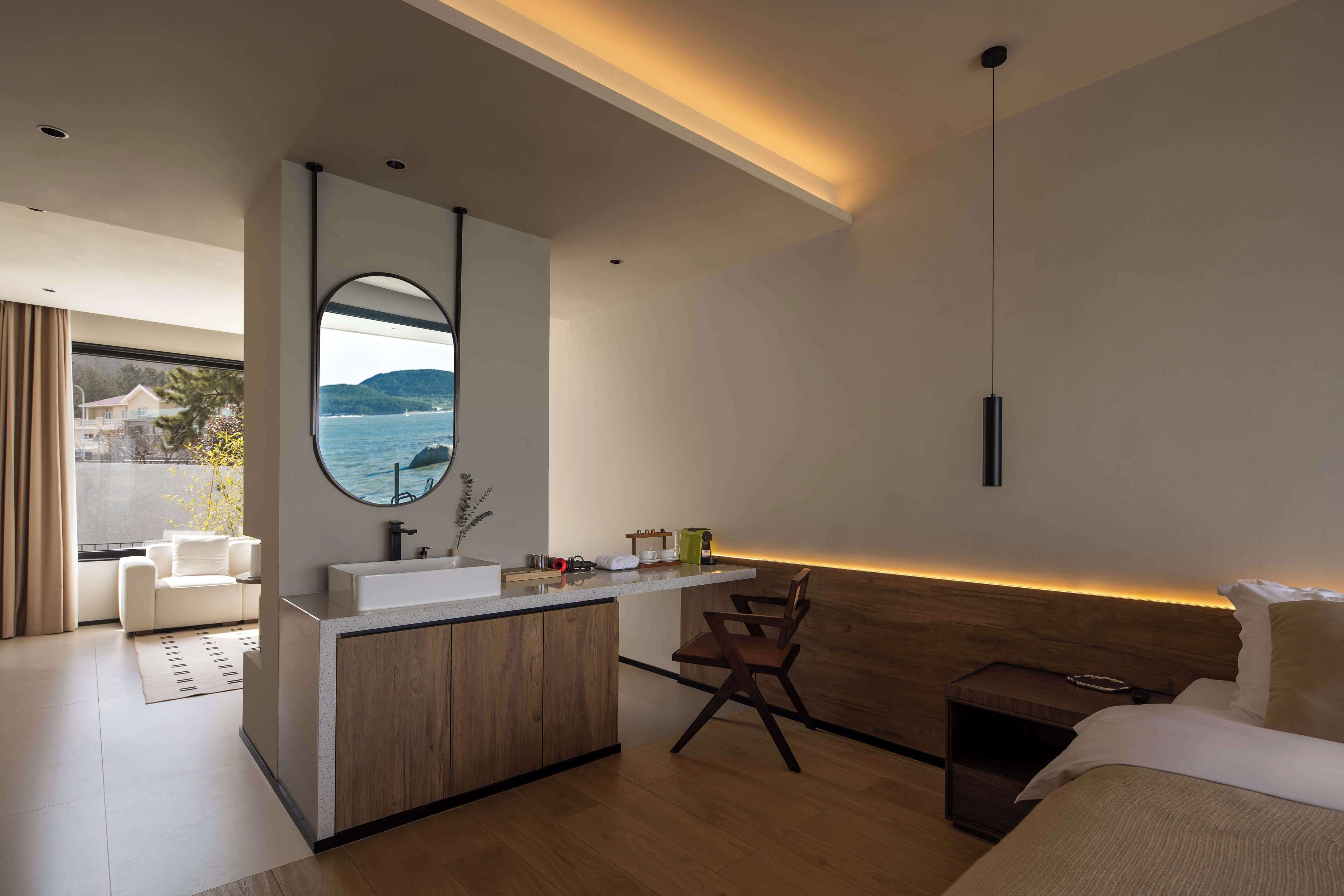A modern bedroom with a wooden headboard, a bedside table, a bed, and a view of the sea through a large window.