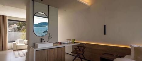 A modern bedroom with a wooden headboard, a bedside table, a bed, and a view of the sea through a large window.
