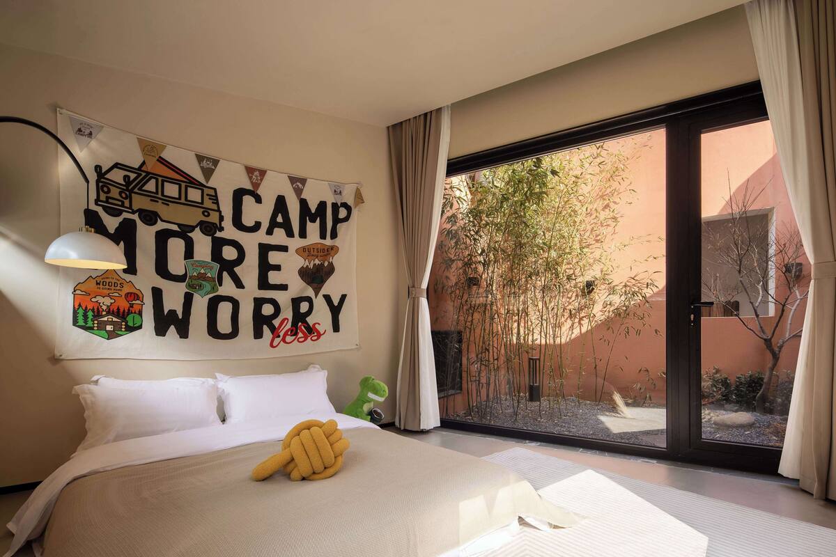 Un dormitorio con una cama grande, una ventana con cortinas, un cartel en la pared y un pequeño dinosaurio de juguete verde.
