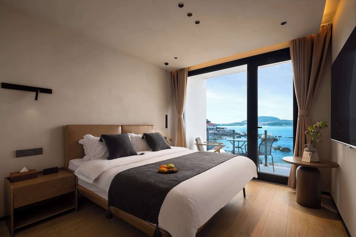 Una habitación de hotel moderna con una cama grande, una mesita de noche, una pequeña mesa con un florero y vista al mar.