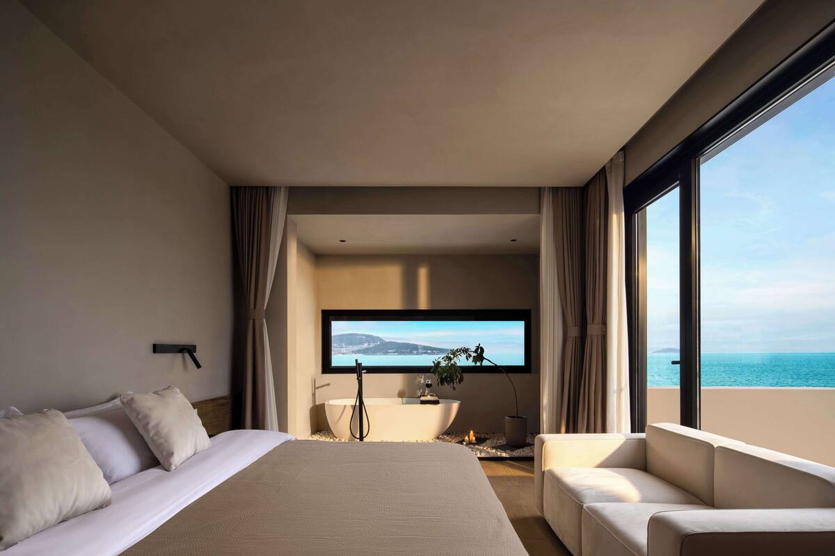 Una habitación de hotel moderna con una cama grande, una zona de estar, una bañera y un ventanal con vistas al mar.