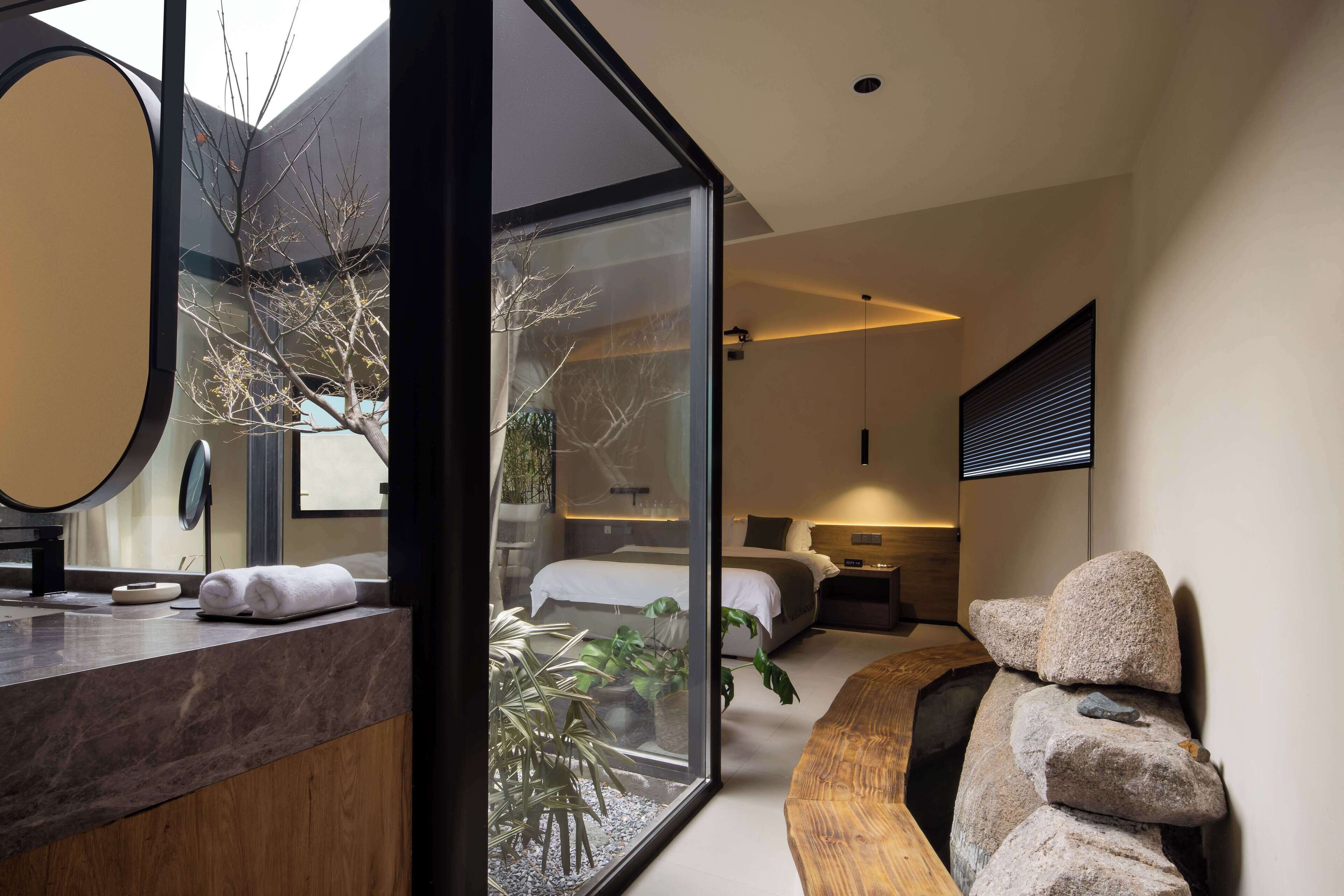 A modern hotel room with a glass wall, a wooden vanity, a bed, and a large mirror.