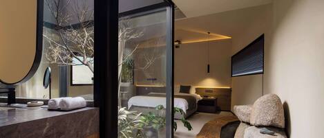 A modern hotel room with a glass wall, a wooden vanity, a bed, and a large mirror.