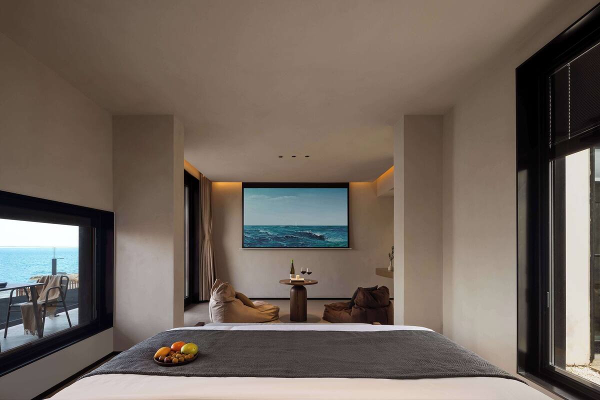 Una habitación de hotel moderna con una cama grande, una mesita redonda pequeña con una botella y vasos, y vista al océano a través de un ventanal amplio.