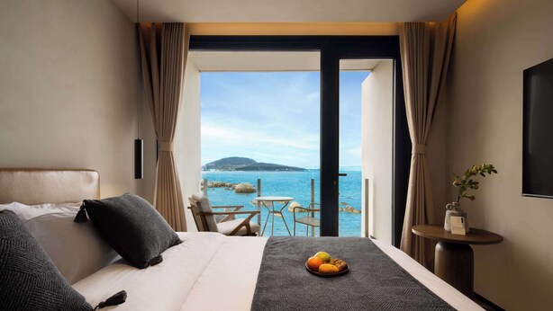 Habitación de hotel con cama, una pequeña mesa con fruta, una silla y vista al mar.