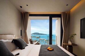 Habitación de hotel con cama, una pequeña mesa con fruta, una silla y vista al mar.