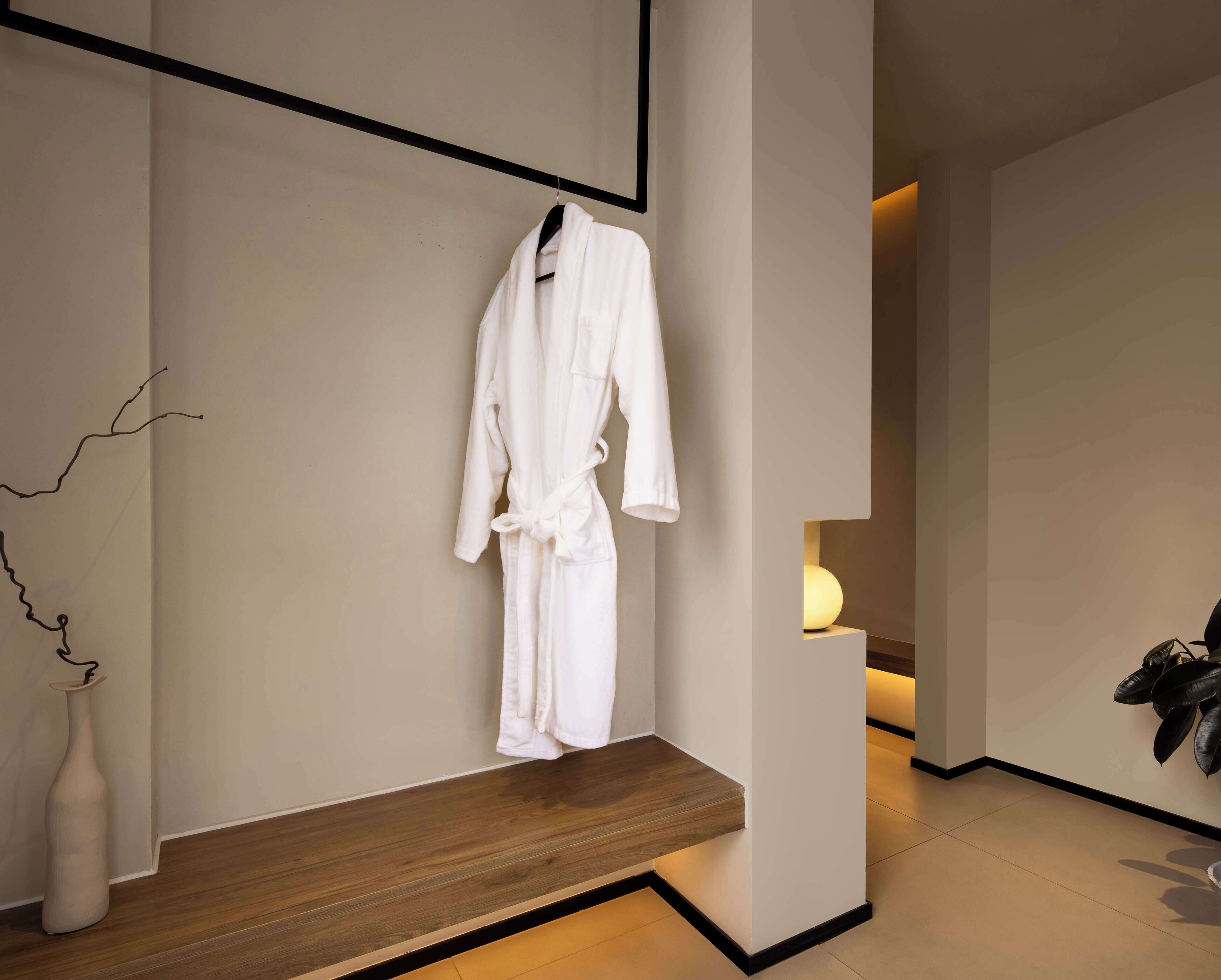 A modern bathroom with a wooden bench, a white robe hanging on the wall, and a vase on the floor.