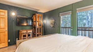 Ein Schlafzimmer mit einem Bett, einem an der Wand montierten Fernseher, einem Holzschrank und einem Fenster mit Blick auf Bäume.