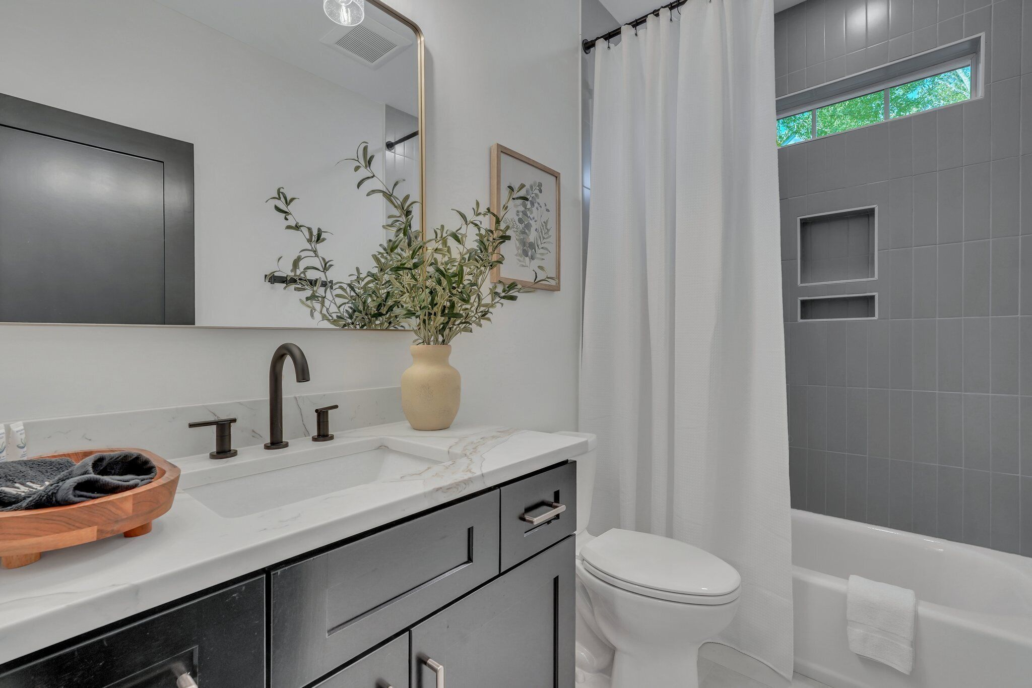 A modern bathroom with a white sink, dark-toned cabinets, a beige vase with a plant, a large mirror, and a bathtub.
