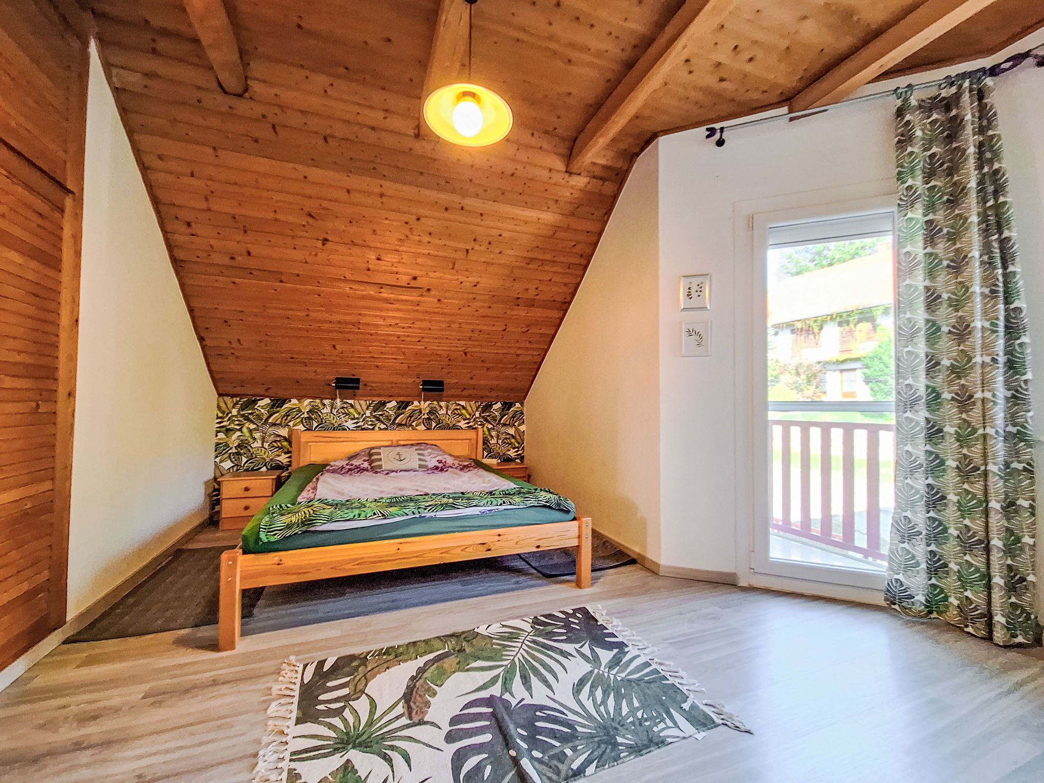 Un dormitorio con techo de madera, una cama con colcha estampada, una mesita de noche de madera, una alfombra pequeña y una ventana con cortinas.