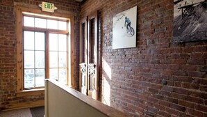 A brick-walled hallway with a large window, an exit sign, and a framed picture of a cyclist. - The Leadville Bunkhouse (Leadville)