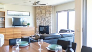 A modern living room with a stone fireplace, a flat-screen TV, and a dining area with a wooden table and chairs. - 94 Aldebaran Rd.  (Ocean Grove)