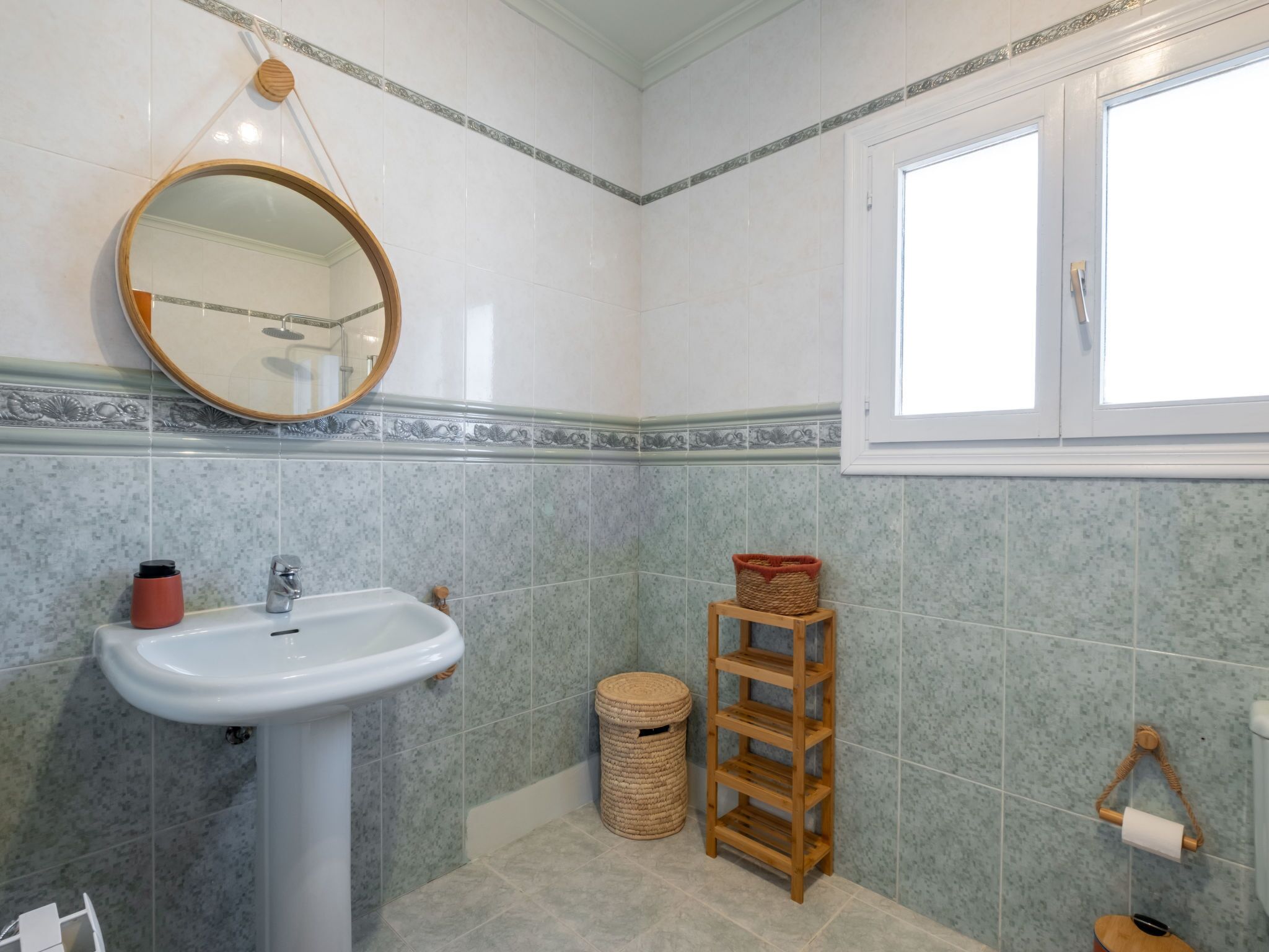 Een badkamer met een witte wastafel, een ronde spiegel, een raam en een houten plank.