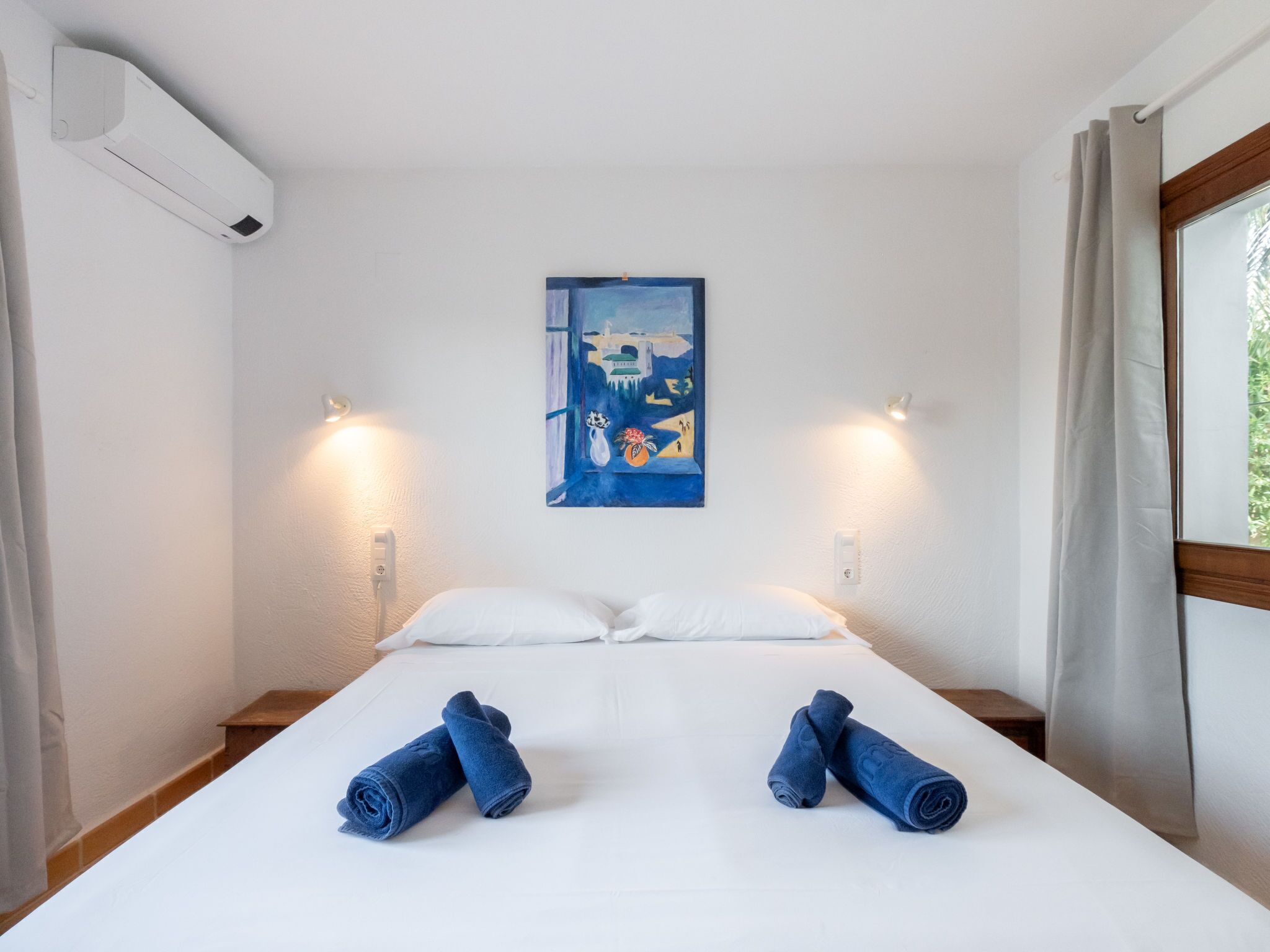 Habitación de hotel con cama, dos lámparas de pared, un cuadro y toallas enrolladas sobre la cama.