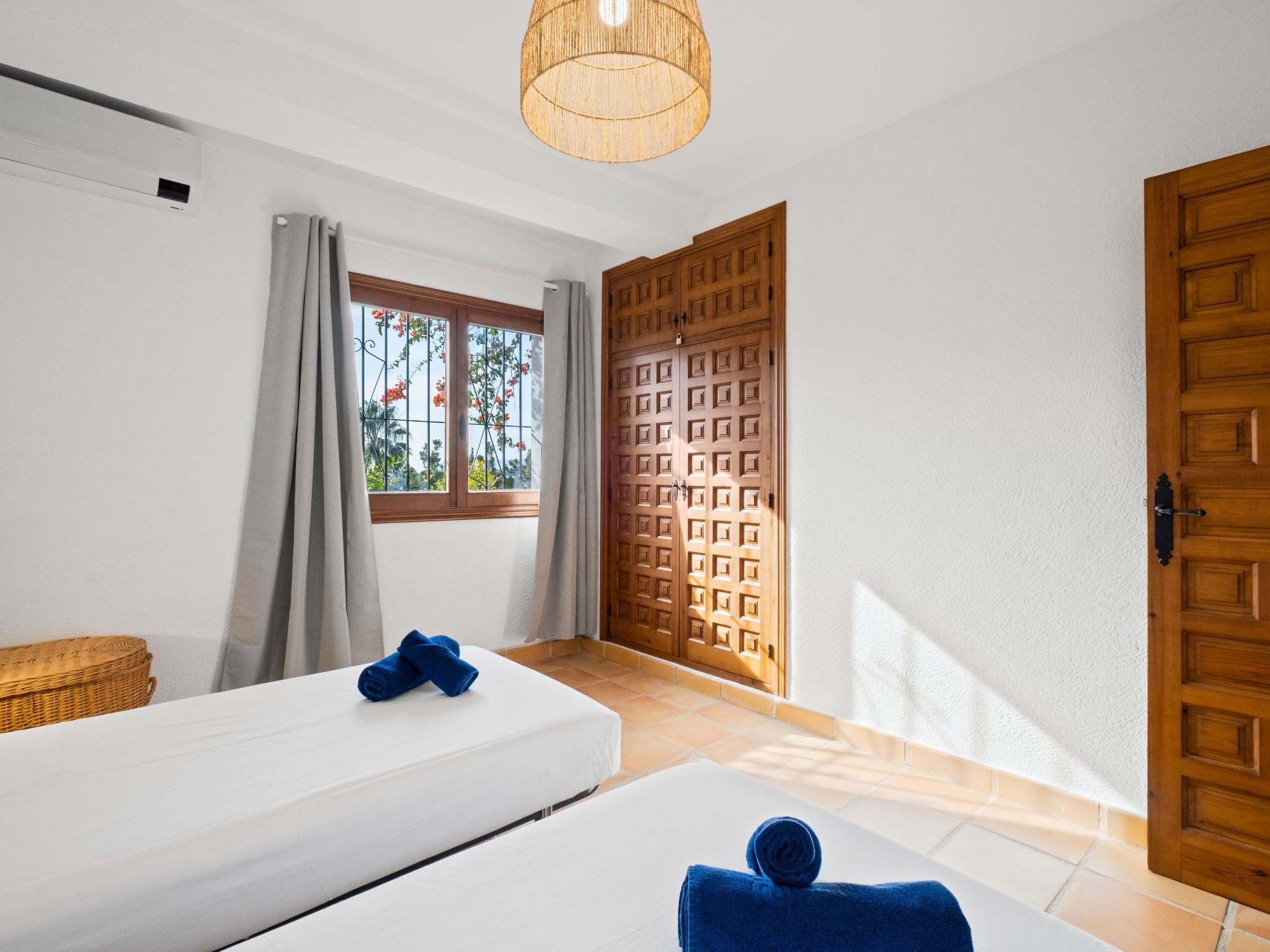 A hotel room with two beds, a window with curtains, a wooden door, and a ceiling light fixture.