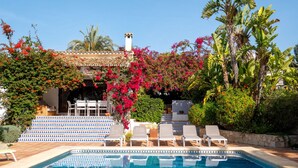 Zona de piscina amb una piscina blava i transparent, gandules blanques i una escala que condueix a una zona d'estar coberta amb un teulat emblumat.